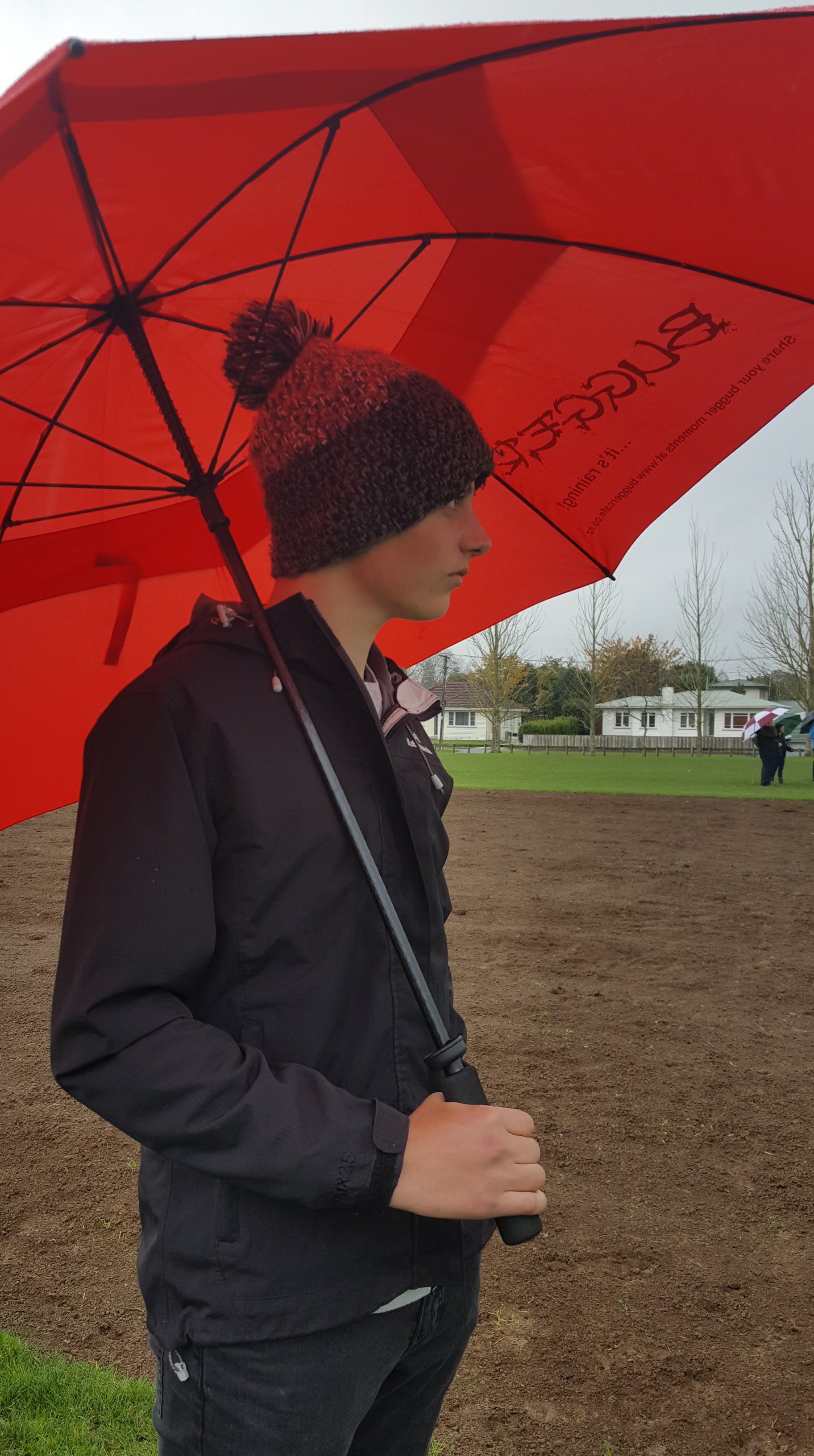Boy wearing a black rain jacket and a woolly hat standing under a red umbrella